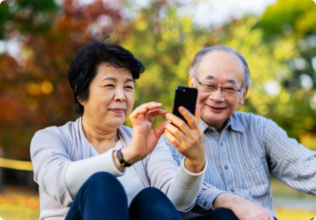 屋外の公園でスマートフォンを操作するシニア夫婦。リラックスした笑顔と親密な様子を表現